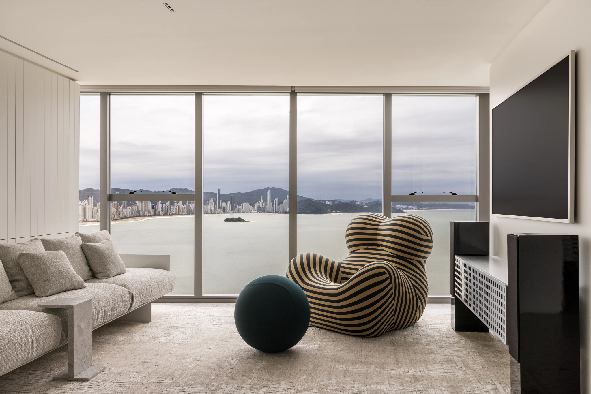 Projeto: Paola Granzotto | Vista da sala de estar com sofá de linhas precisas de Jader Almeida e poltrona Up, com obras de arte nas paredes e vista para o mar.